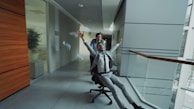 Man in suit rides office chair pushed by colleague
