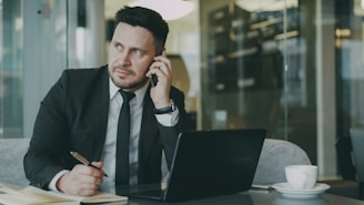 Man in suit talking on phone at desk with laptop.