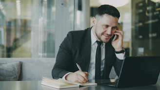Man in suit talking on phone while writing in notebook.