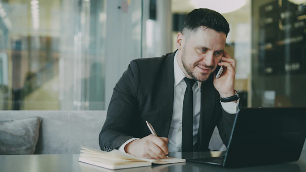 Man in suit talking on phone while writing in notebook.