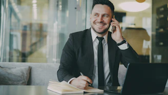 Man in suit talking on phone at desk with laptop.