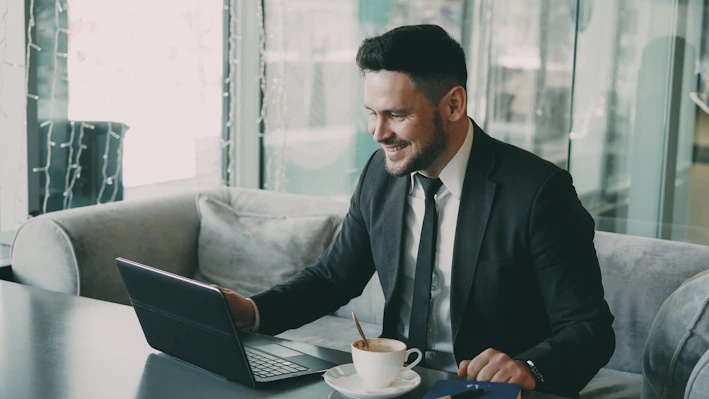 Man in suit smiles while working on laptop with coffee.