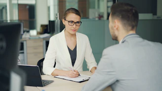 Woman in glasses interviews man at office desk.