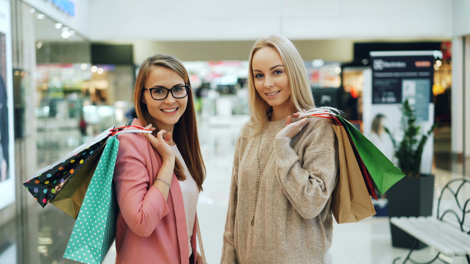 Two smiling women holding shopping bags in a mall.