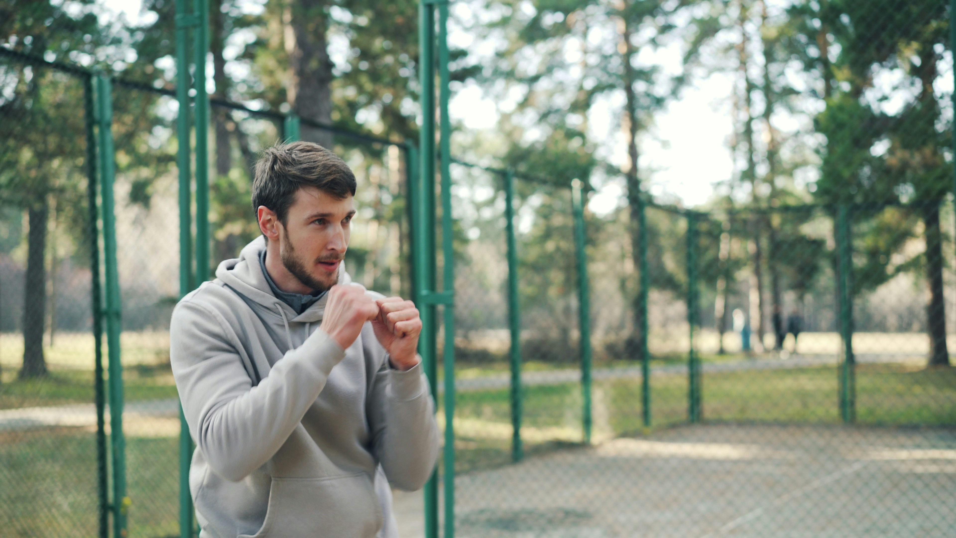 Man in hoodie boxing outdoors near fence.