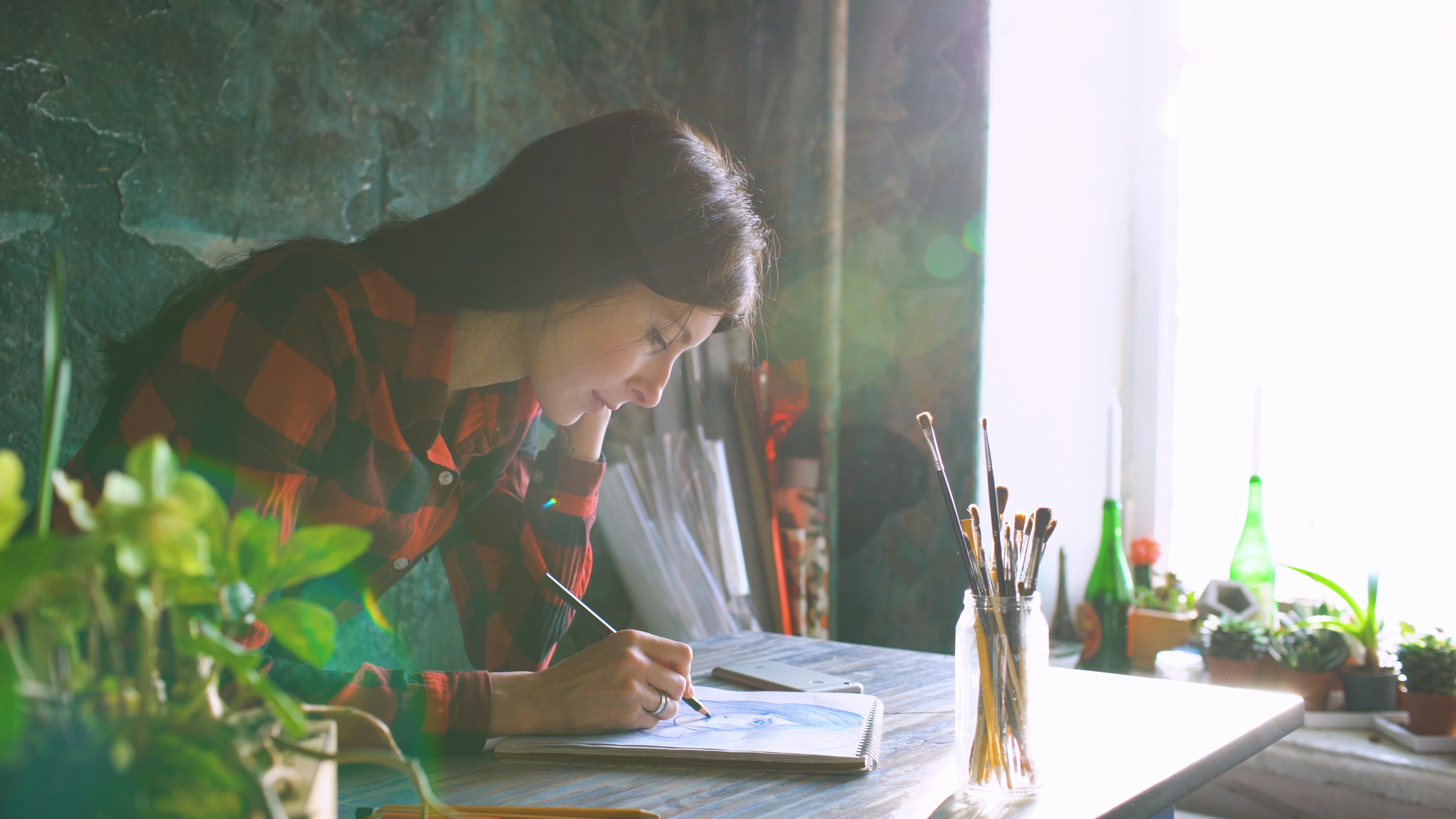 Young woman artist painting sketch on paper notebook with pencil in studio indoors. Flecks of sunlight