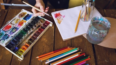Artist painting with watercolors and colored pencils on wooden table.