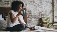 Woman drinking coffee while using laptop in bed.