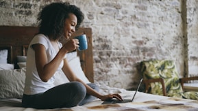 Woman drinking coffee while using laptop in bed.