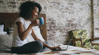 Woman drinking coffee while using laptop in bed.