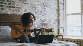 Young woman playing guitar on bed with laptop.