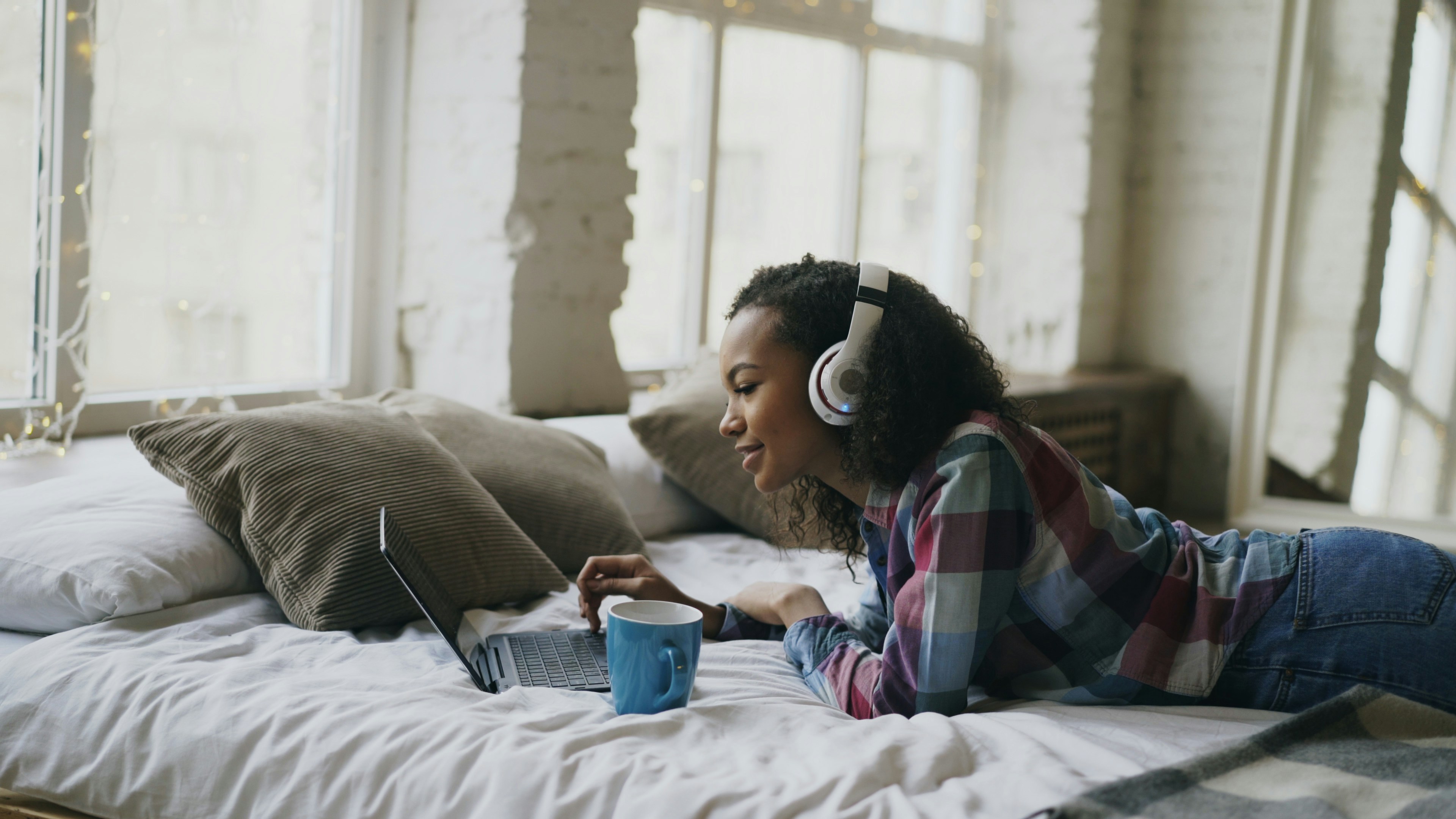 Attractive mixed race girl listen to music while surfing social media on laptop lying on bed at home