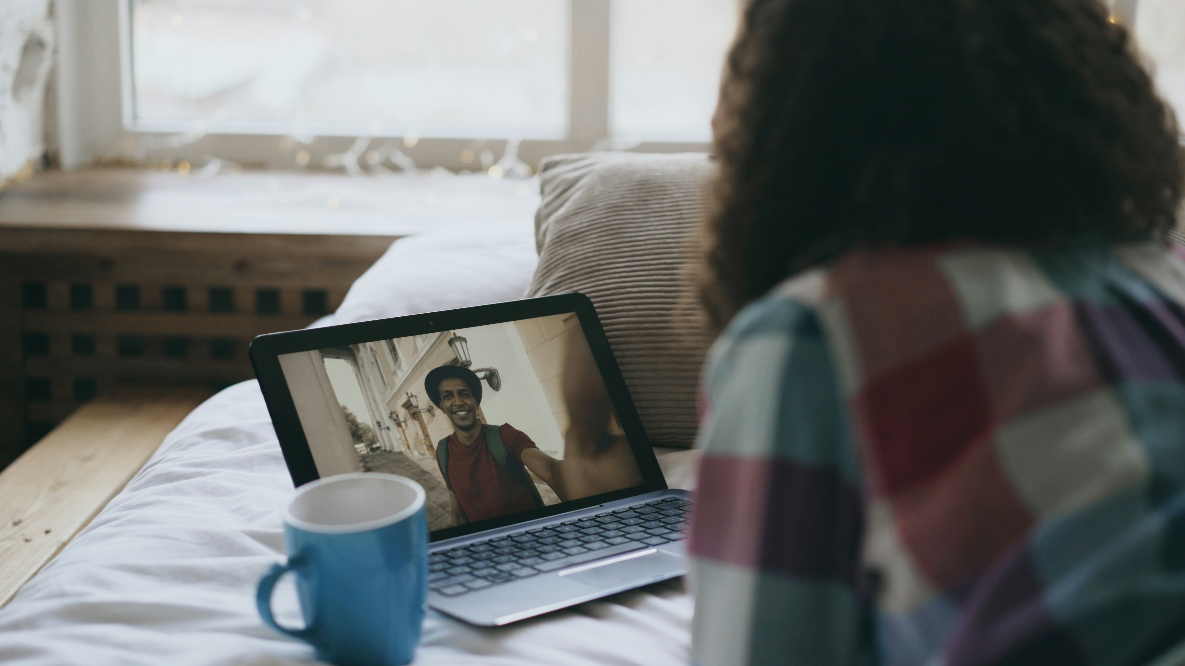 Curly young woman having online video chat with friend using laptop camera while lying on bed at home