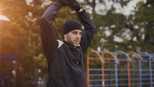 Man in black hat stretching outdoors with arms raised outdoors.