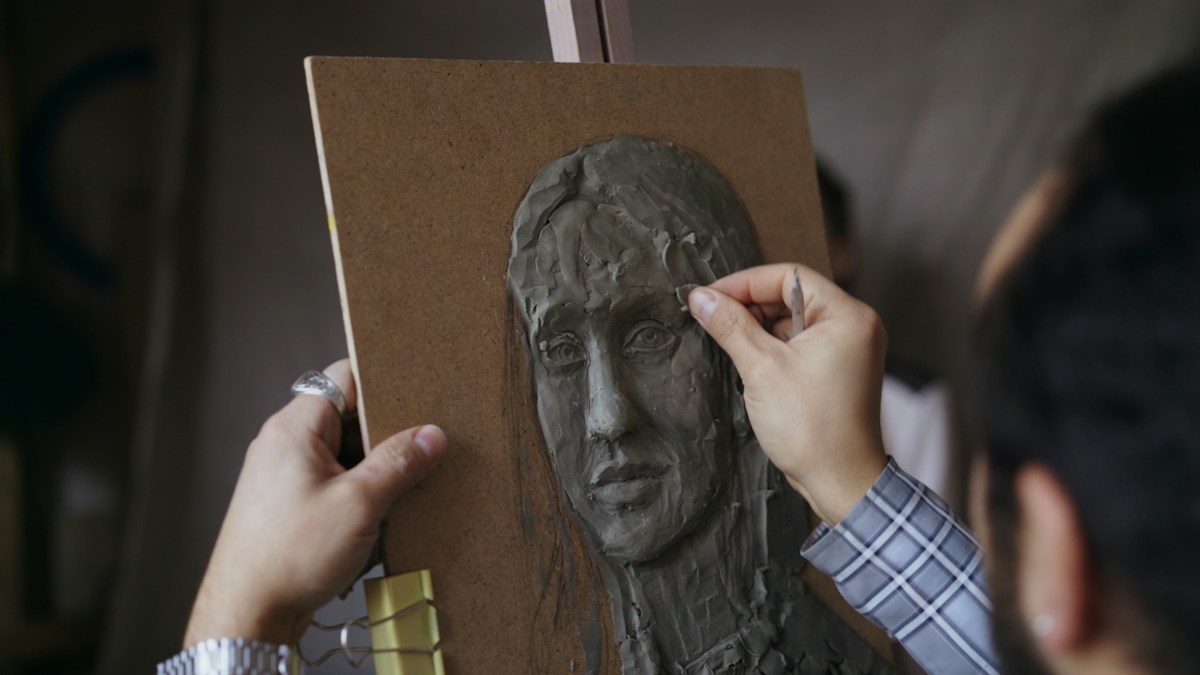 Un sculpteur façonne un visage en argile grise sur un panneau posé sur un chevalet
