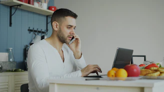 Man talking on phone while working on laptop at table.