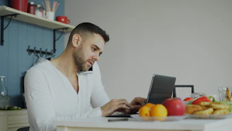 Man on phone and laptop at kitchen table.