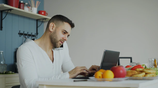Man on phone and laptop at kitchen table.