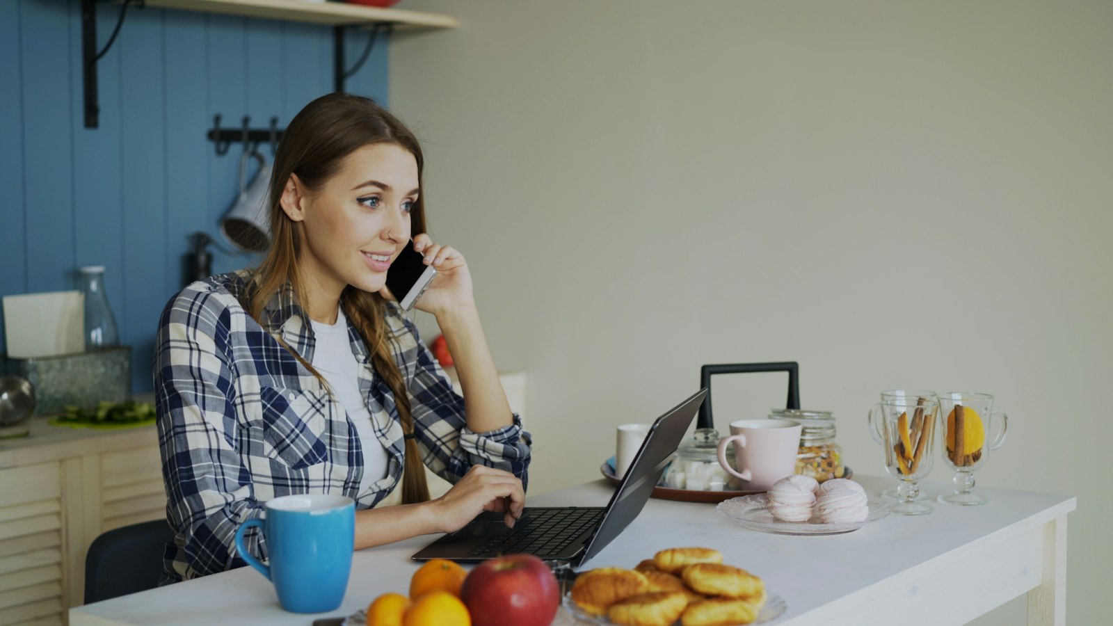 Une femme en chemise à carreaux téléphone dans sa cuisine, son ordinateur portable ouvert sur la table à côté d'une tasse bleue et de fruits.