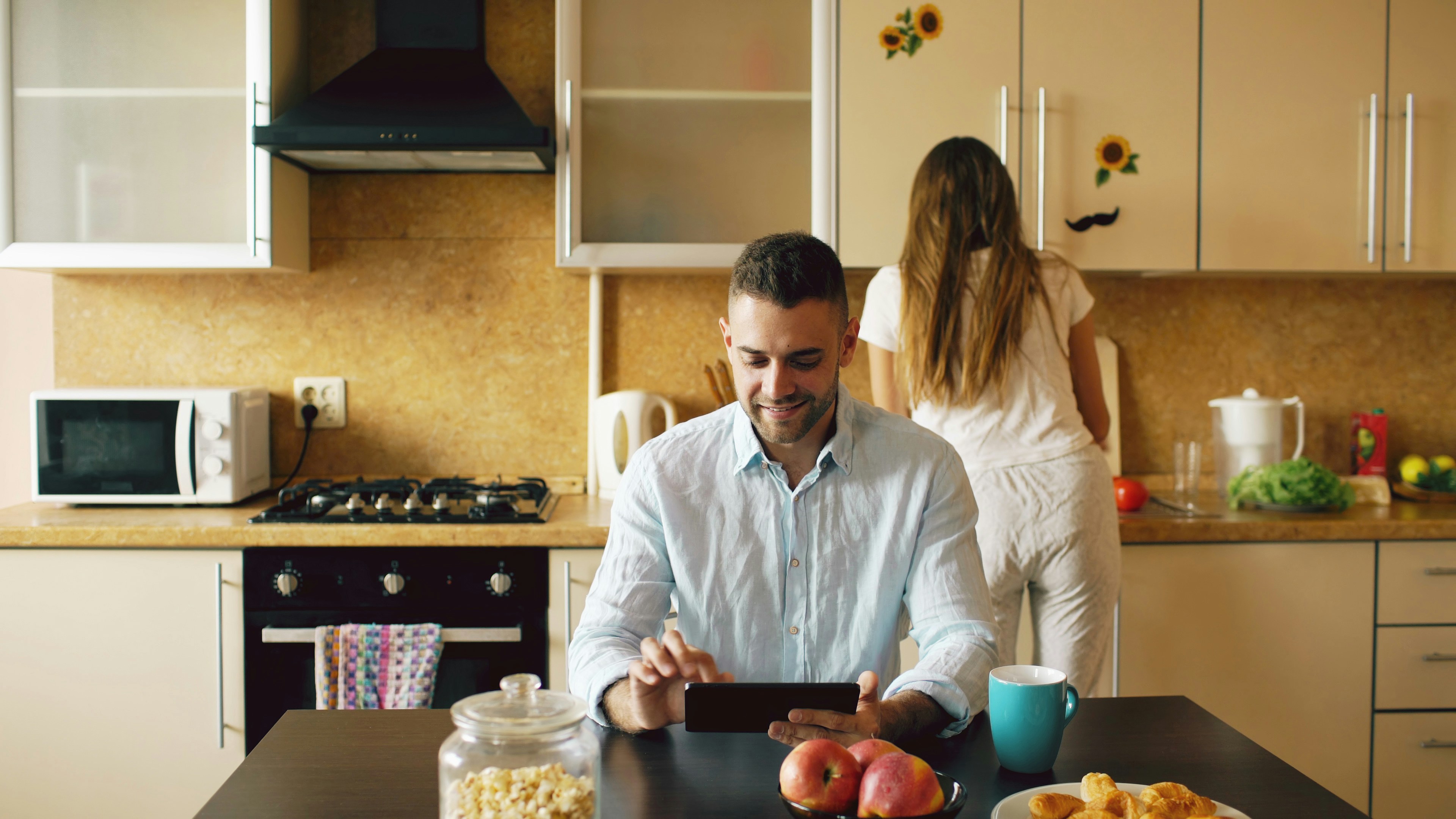 Couple in kitchen, man using tablet at table.