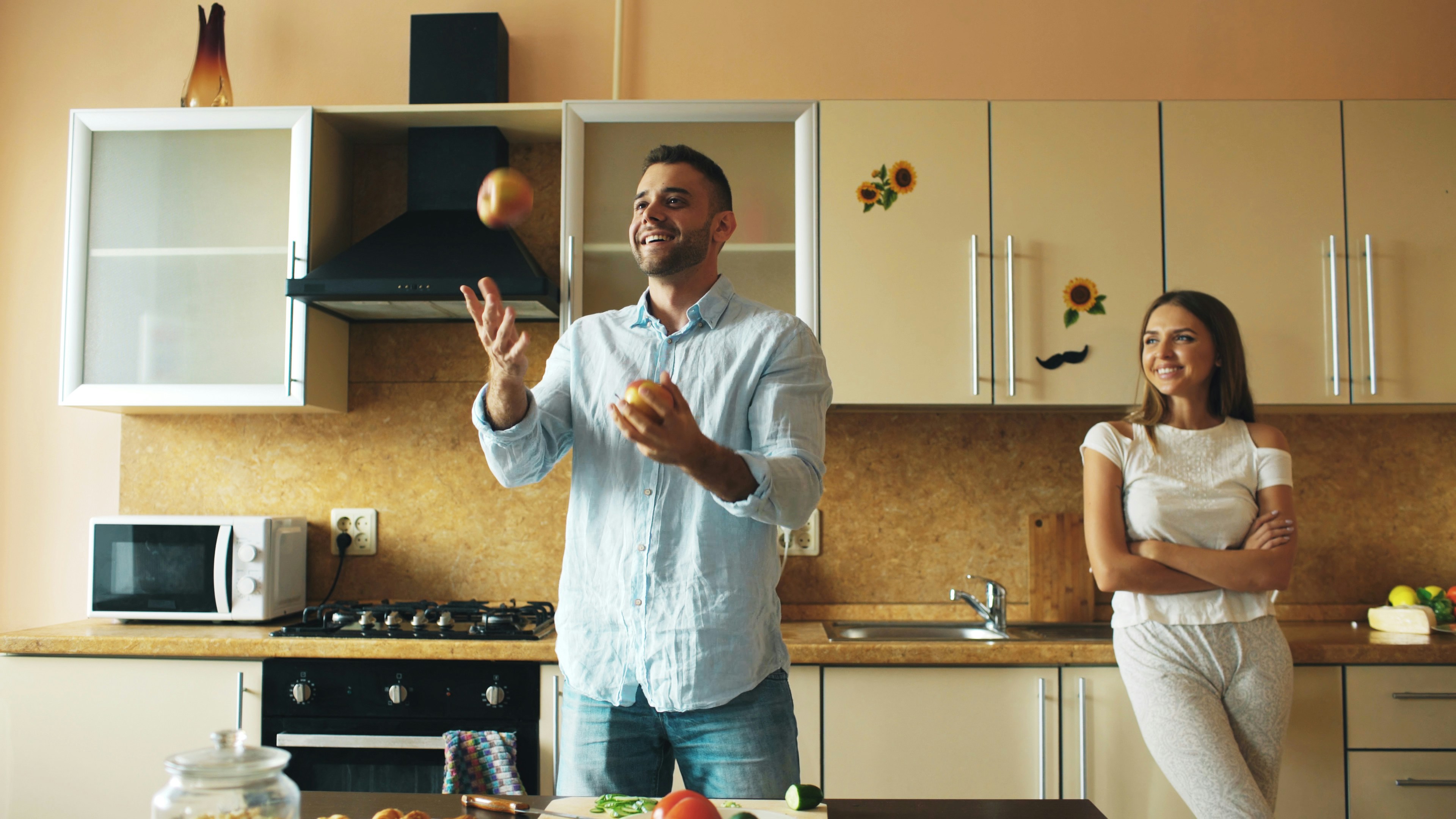 Man juggles fruit while woman watches in kitchen.