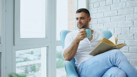 Man drinking coffee and reading book by window.