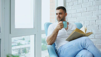 Man drinking coffee and reading book by window.