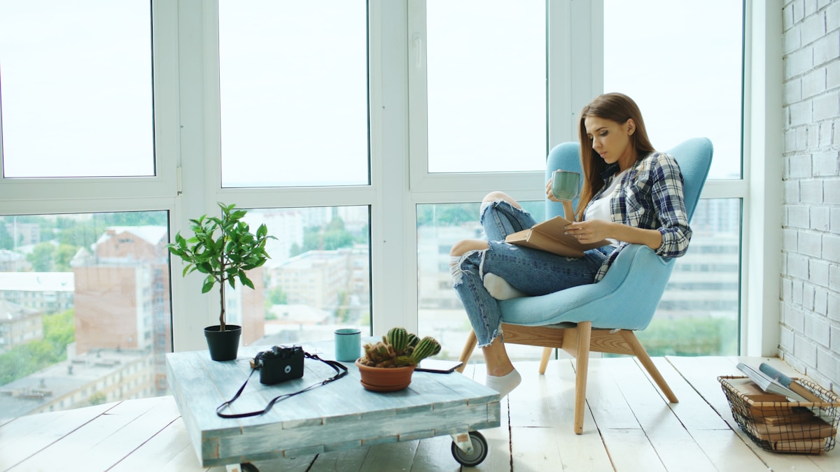 woman reading a book in a blue armchair by large apartment windows with plants and a pallet coffee table