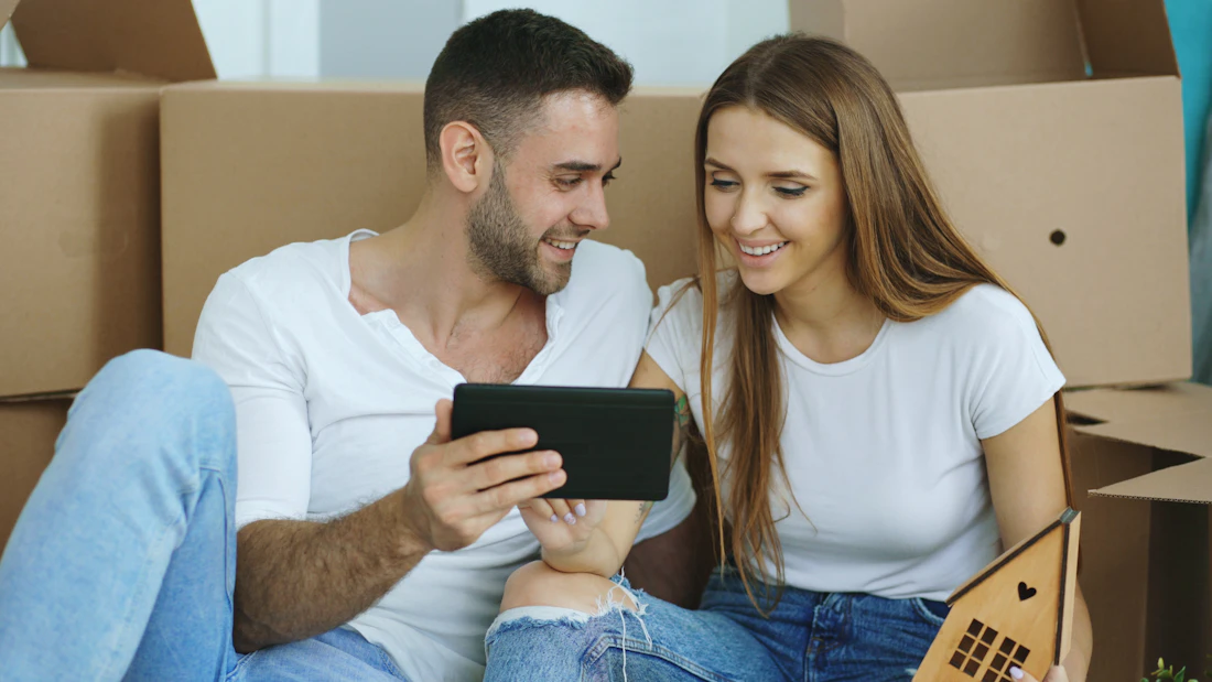Pareja sonriendo mientras revisa opciones en una tablet, rodeada de cajas de mudanza en su nuevo departamento