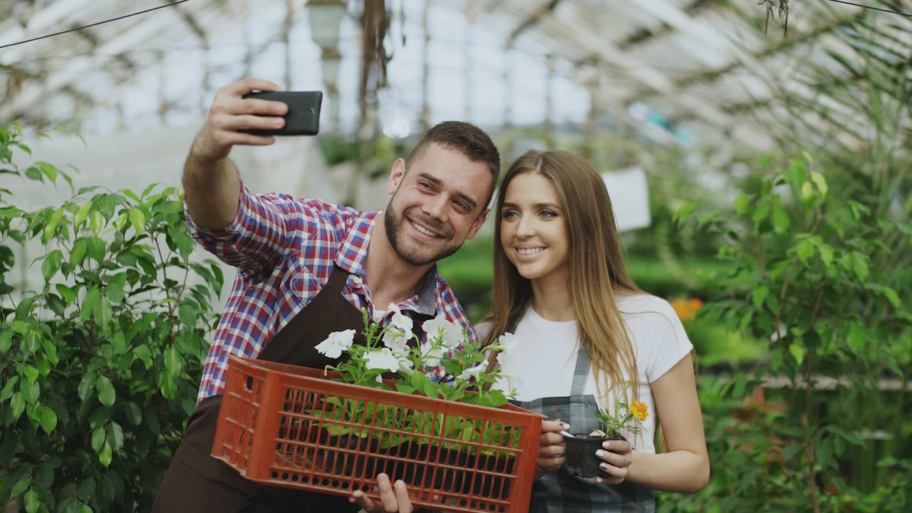 Two growers in aprons smile for a phone selfie in a greenhouse, one holding a crate of white flowers.