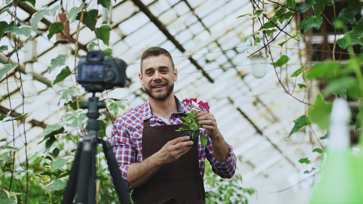 Un homme souriant en chemise à carreaux et tablier marron tient un pot de fleurs roses face à un appareil photo posé sur un trépied, dans une serre pleine de plantes