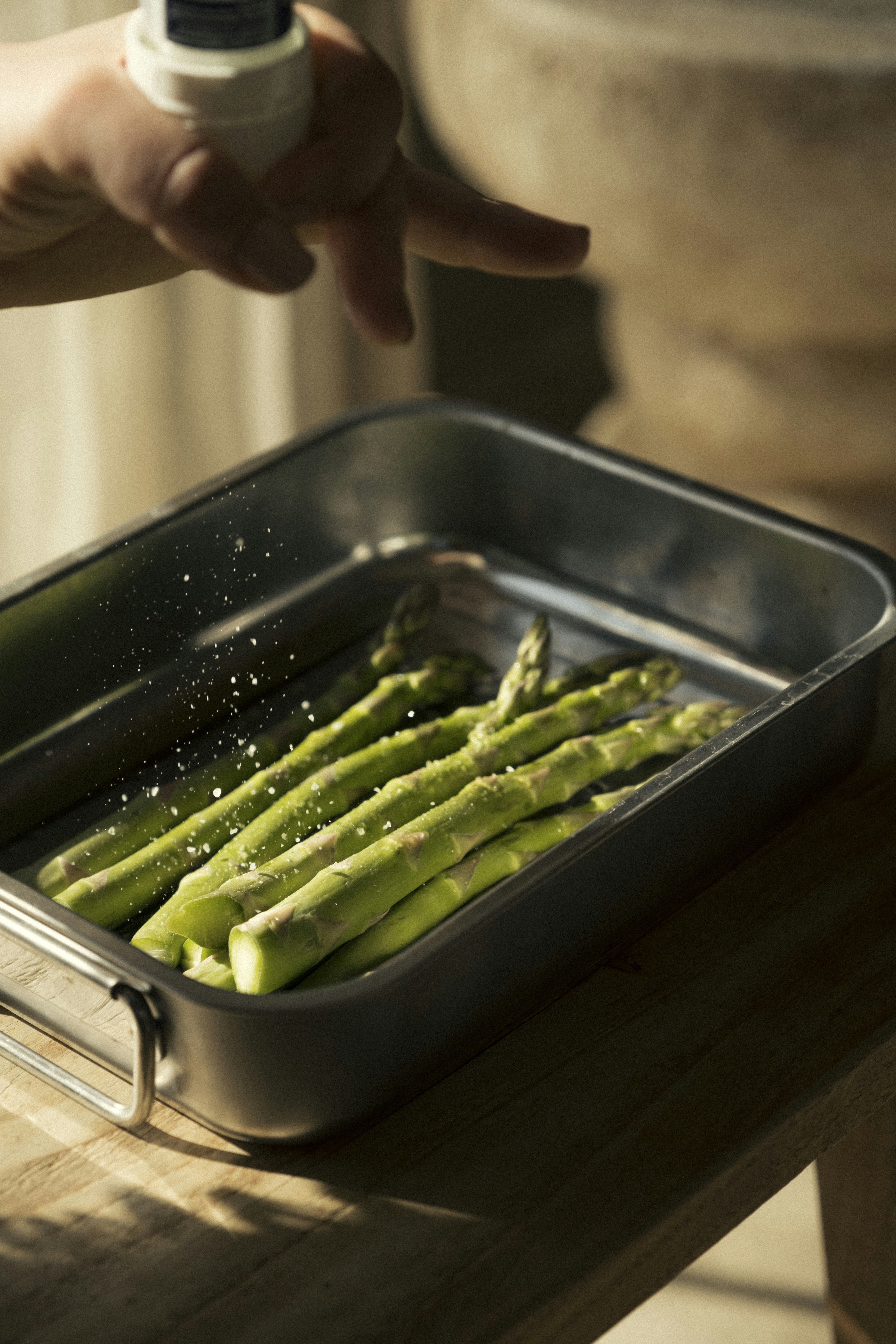Hand seasoning asparagus in a baking dish.