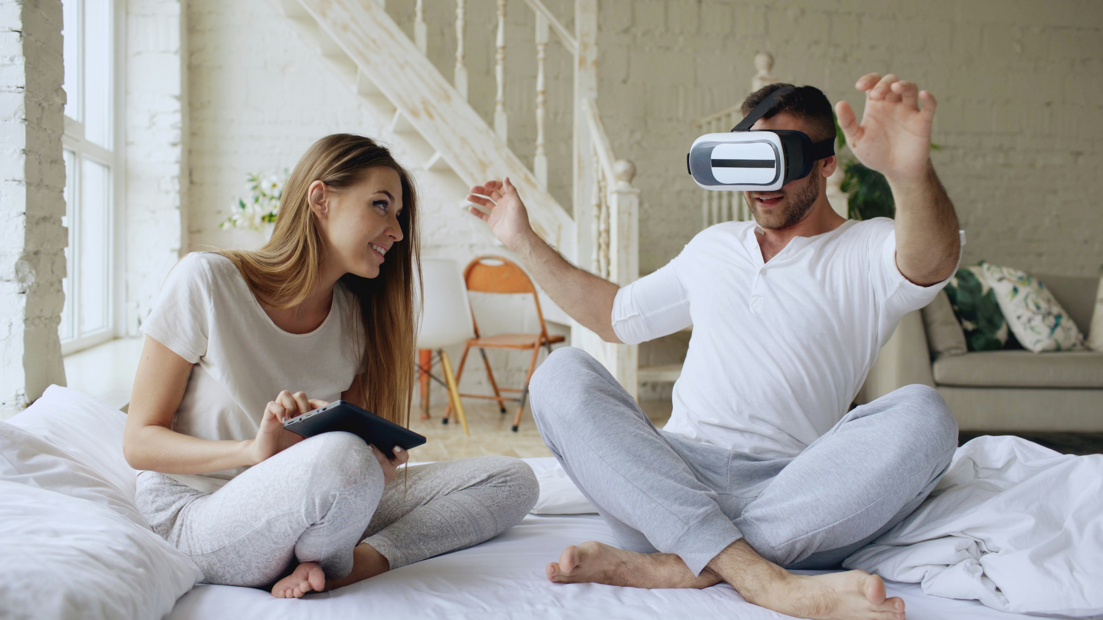 Young cute couple with tablet computer and virtual reality headset playing 360 VR video game while sitting in bed at home. Woman control her boyfriend entertainment using touchpad