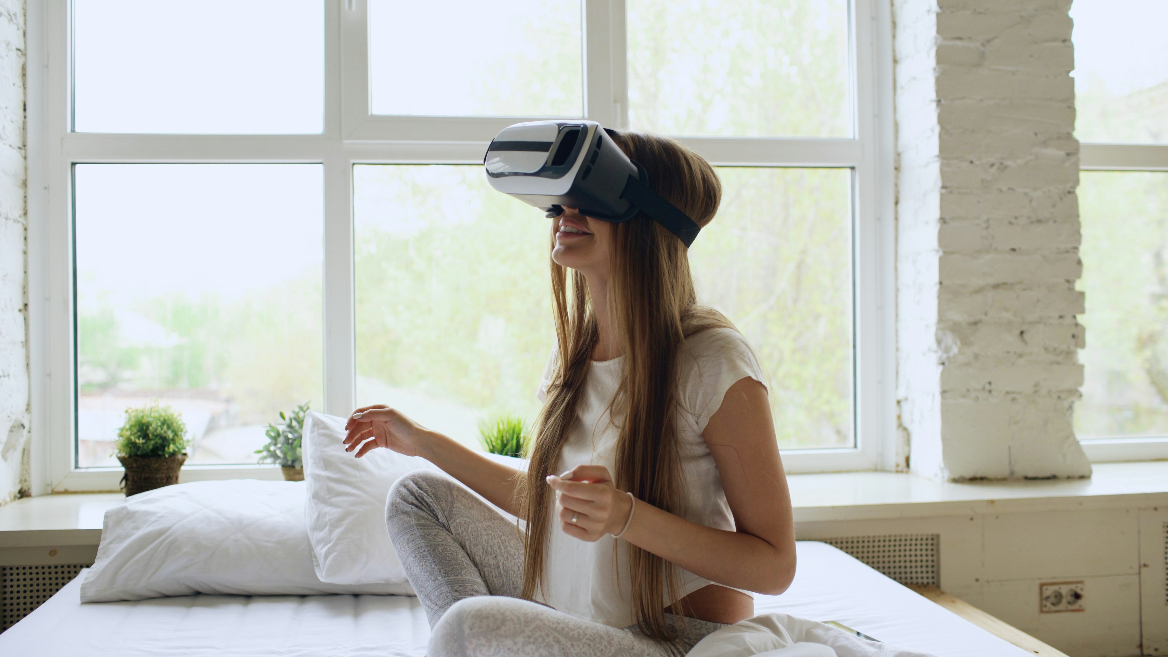 Young cheerful woman wearing virtual reality headset watching 360 VR video movie sitting in bed at home