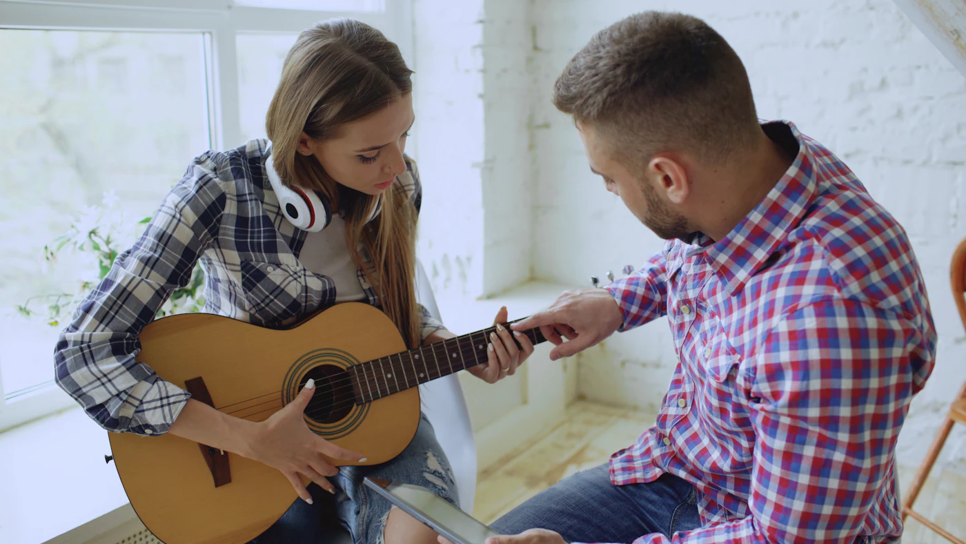 A tutor guiding a student's hand on the fretboard of an acoustic guitar