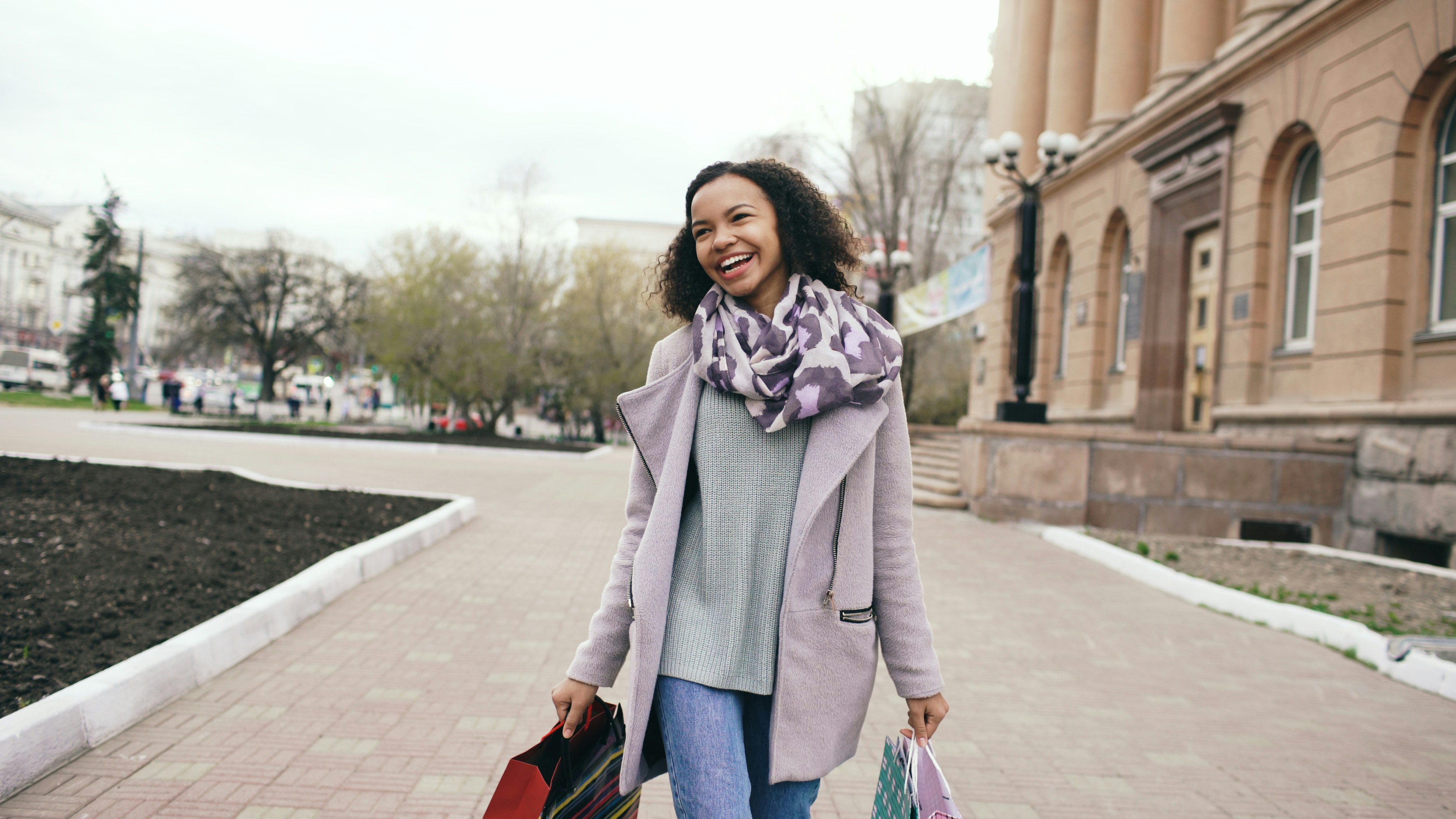 A smiling woman walks down a sidewalk with shopping bags.