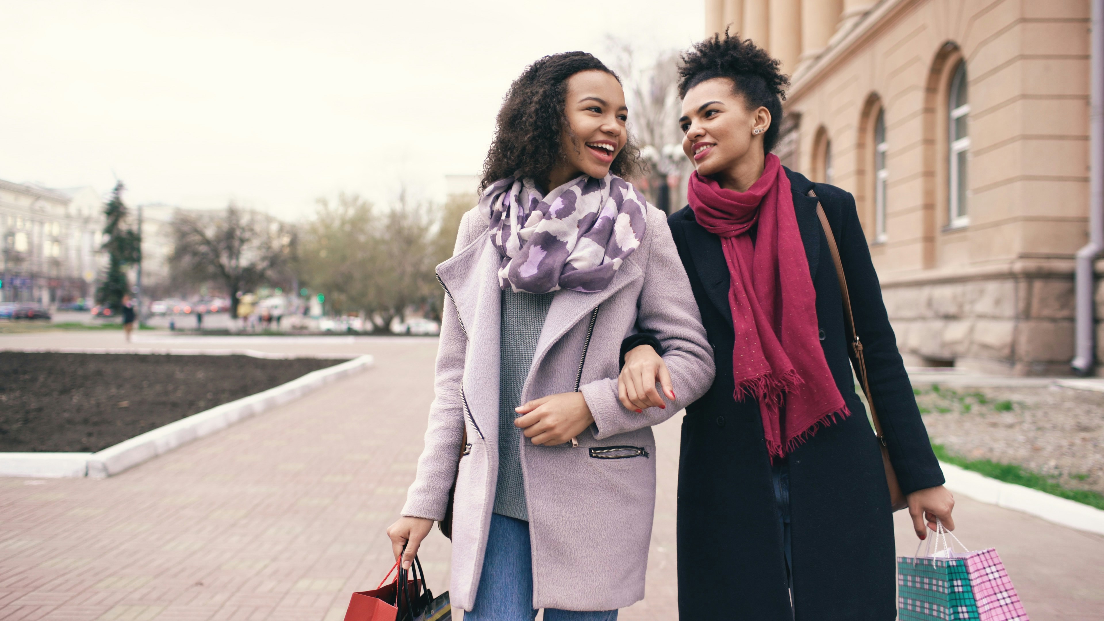 Two women walk arm-in-arm with shopping bags.