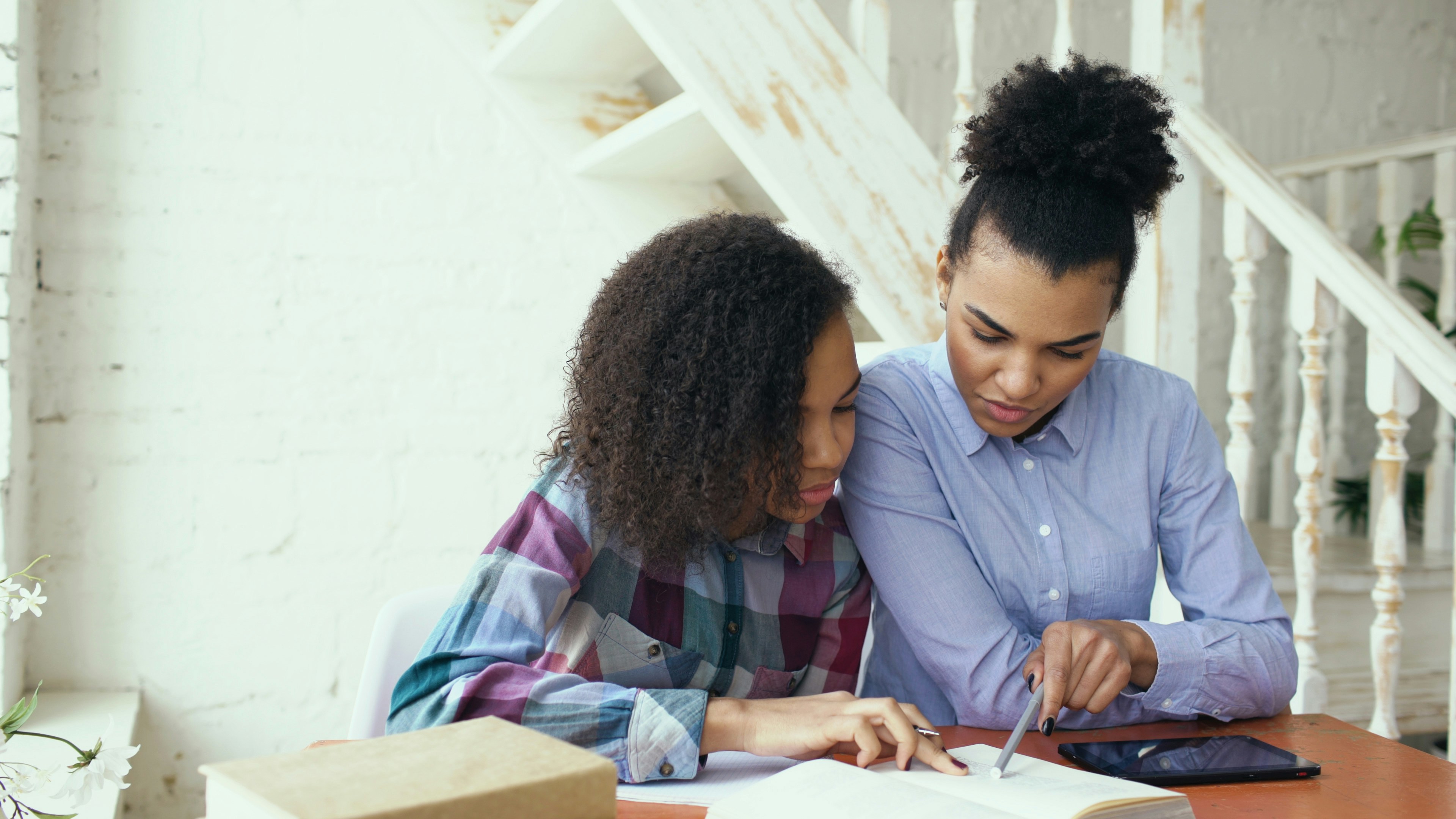 Teenage curly haired mixed race young girl sitting at the table concentrating focused learning lessons and her elder sister helps her studying at home