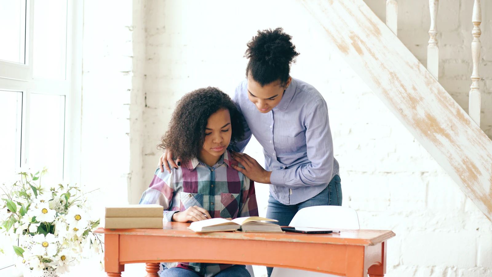 A teenage girl and her older sister sitting together at a table, the older sister helping her with schoolwork.
