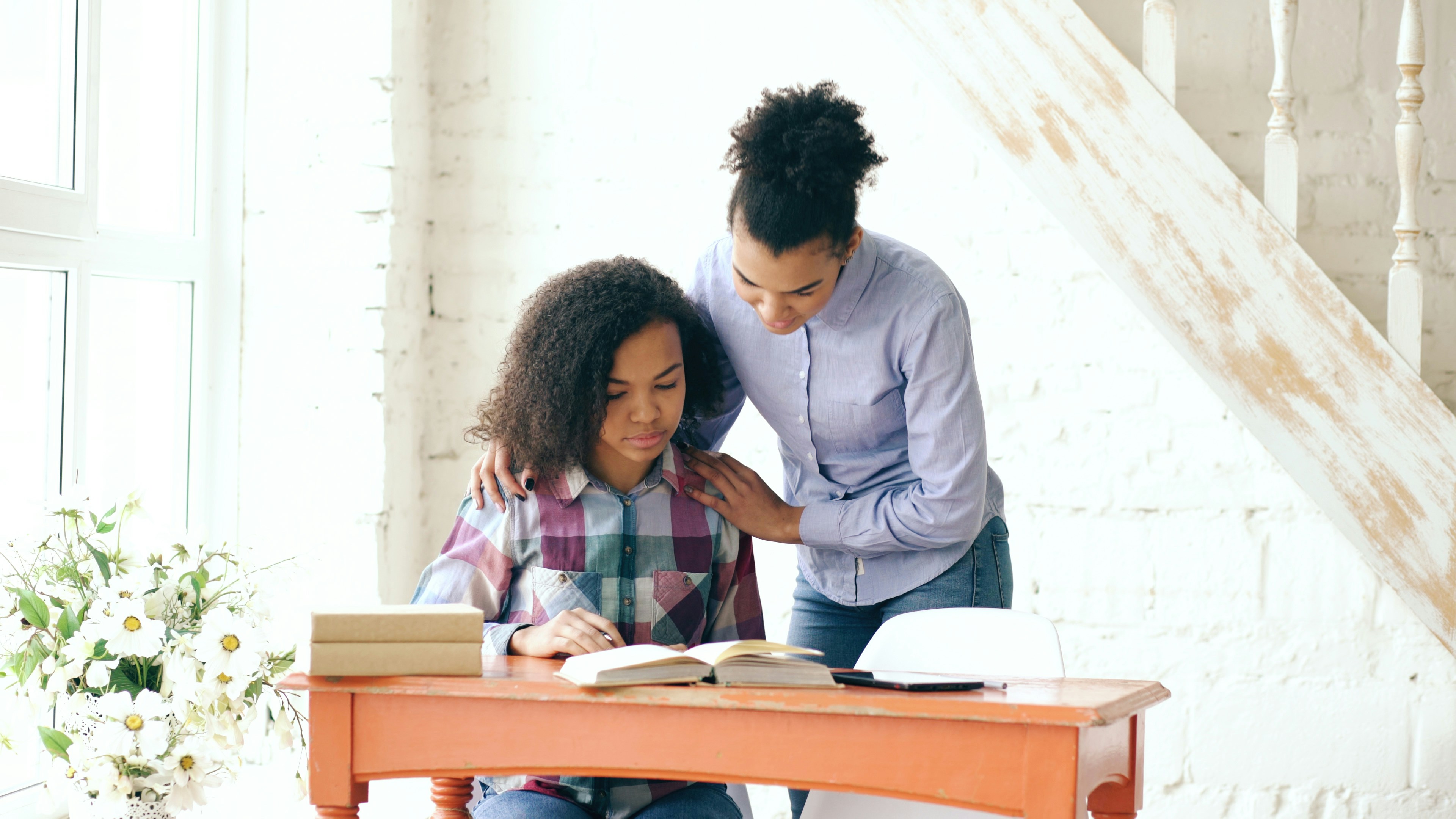 Teenage curly haired mixed race young girl sitting at the table concentrating focused learning lessons and her elder sister helps her studying at home