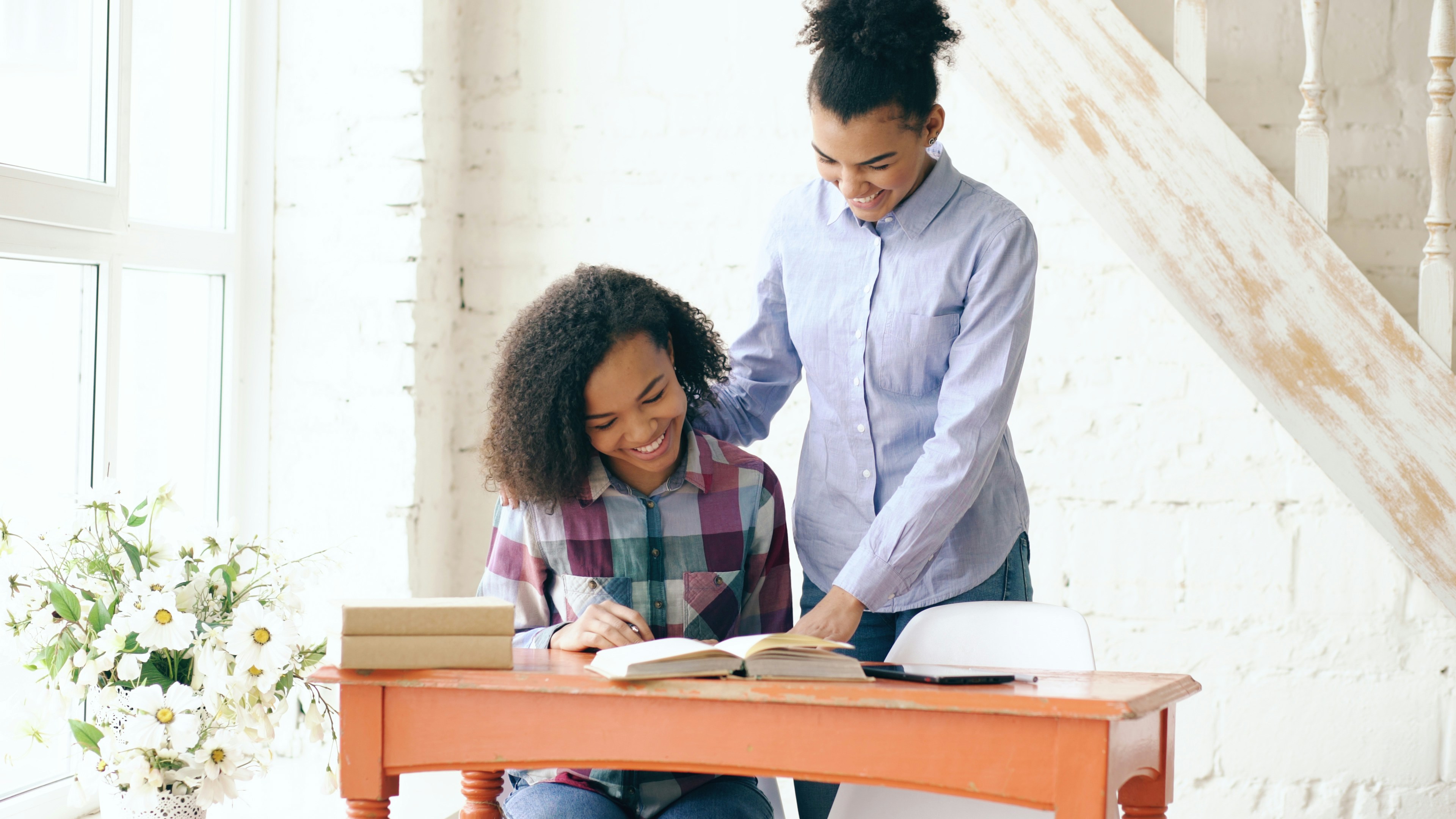 Teenage curly haired mixed race young girl sitting at the table concentrating focused learning lessons and her elder sister helps her studying at home