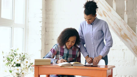 Woman helps girl with homework at desk.