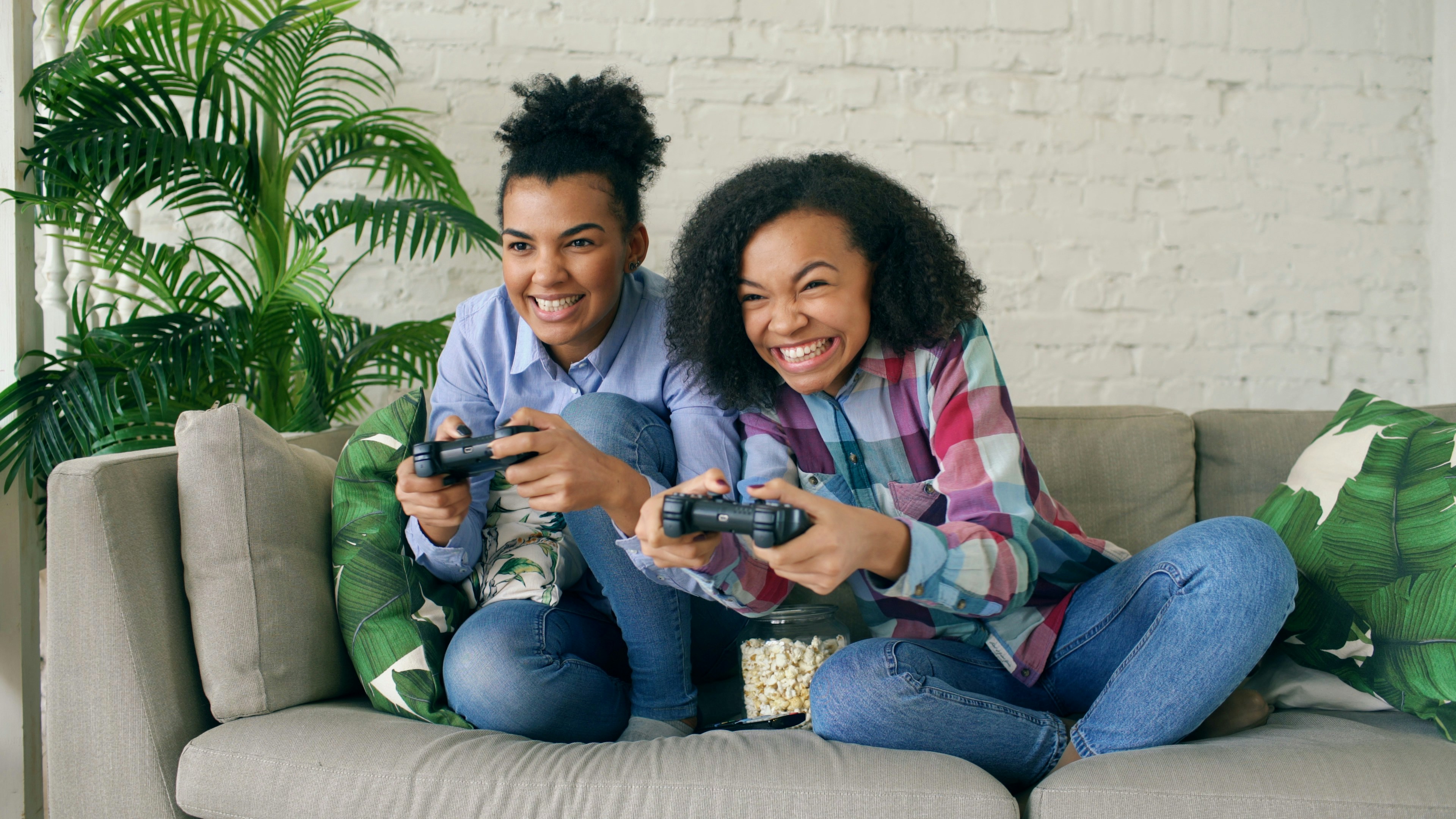 Two mixed race curly girl friends sitting on couch play console computer games with gamepad and have fun