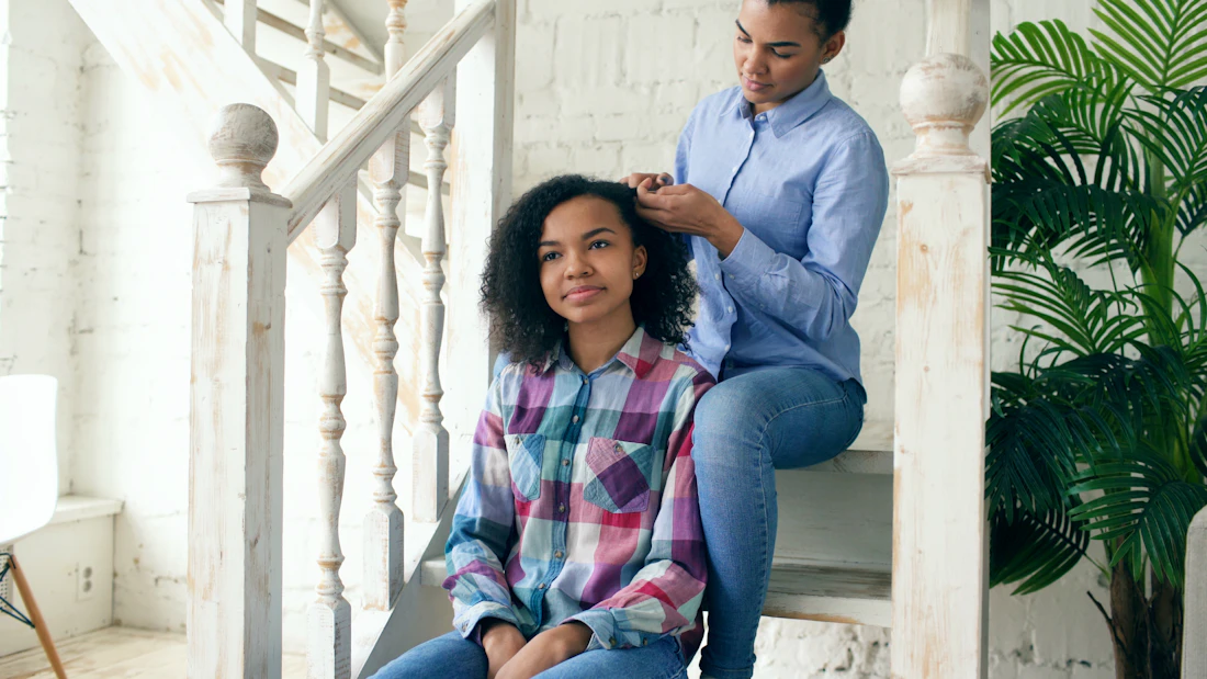 A guardian gently styling her teenage daughter's hair at home as they spend time together
