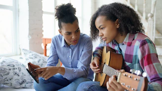 Two young women learning guitar together indoors.