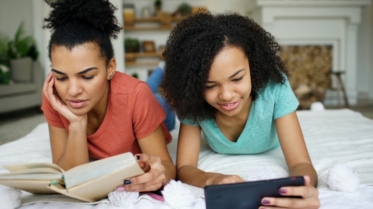 Two teen girls lying on a bed, one reading a book and one using a tablet