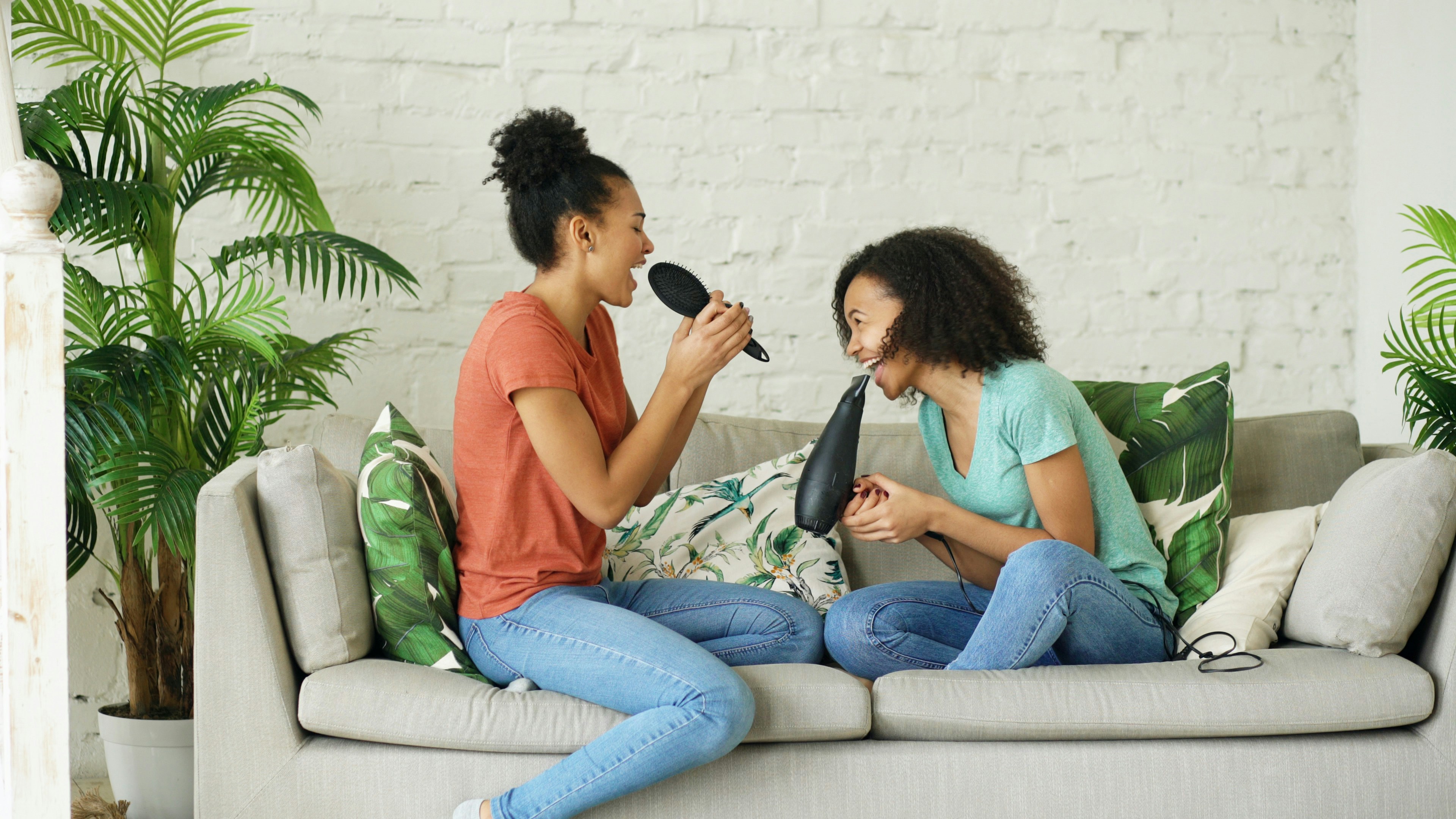 Two women singing into hairbrushes on a couch.
