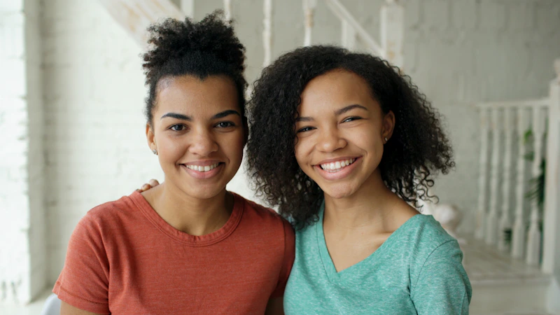 Two smiling young sugar babies standing together indoors.