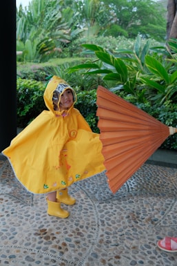 Young child in yellow raincoat holds large orange umbrella