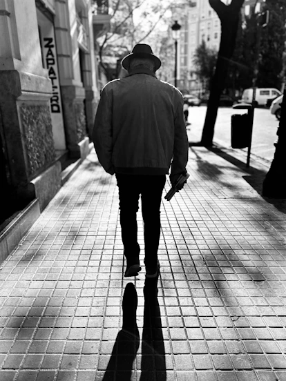 Man in hat walks down sunny sidewalk holding object.